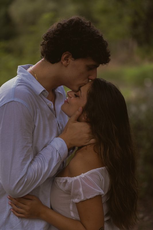 Bacio ravvicinato: lui le tiene il viso tra le mani, vegetazione sfocata sullo sfondo