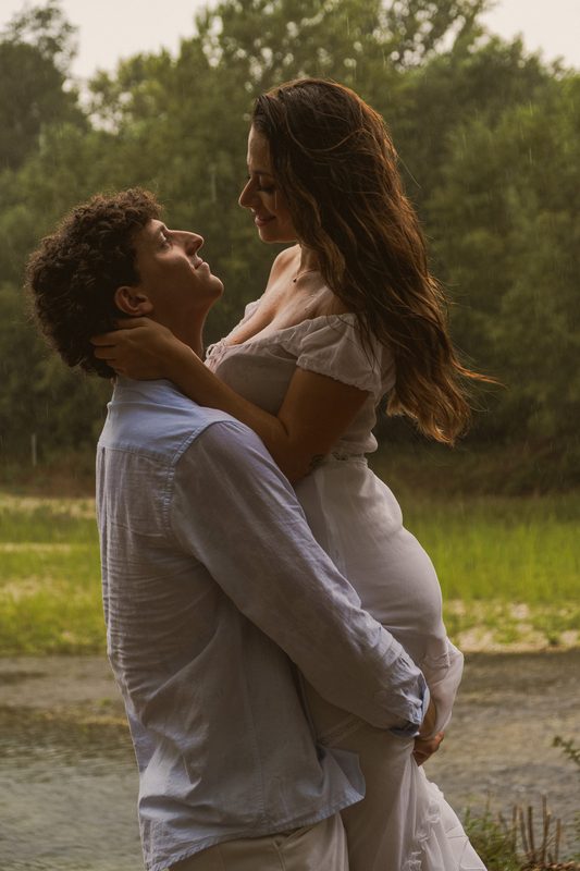 Lui la solleva tenendola stretta, lei sorride guardandolo dall'alto, capelli bagnati e vegetazione dietro