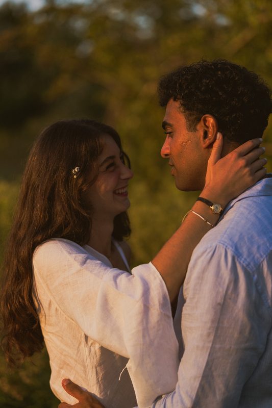 Lei gli cinge il collo con le braccia e ride guardandolo, luce dorata e vegetazione dietro