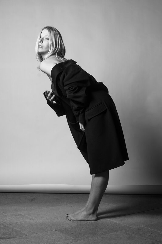 Ritratto in studio: donna con cappotto nero, in piedi piegata in avanti, a piedi nudi su fondo chiaro