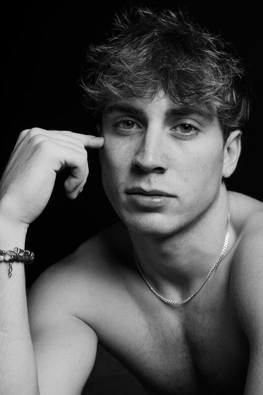 Ritratto in studio ravvicinato: giovane uomo con dito appoggiato alla tempia, collana e bracciali, fondo nero