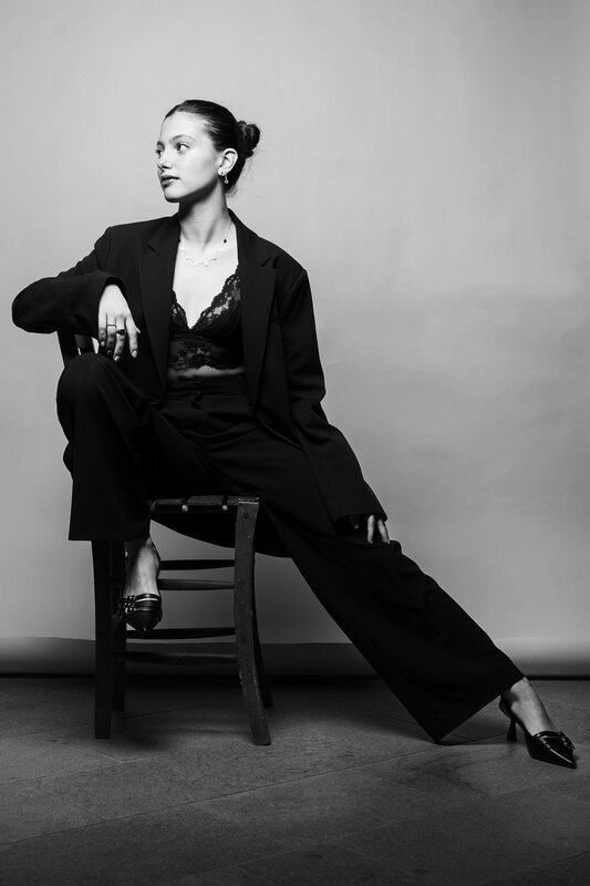 Ritratto in studio: donna in completo nero seduta su una sedia di legno, gambe distese, fondo chiaro