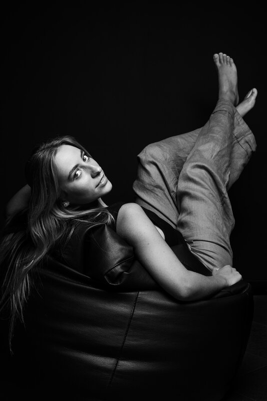 Ritratto in studio: donna distesa su un pouf di pelle con le gambe sollevate, fondo nero