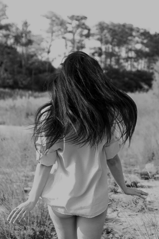 Bianco e nero: ragazza di spalle con i capelli al vento davanti a una pineta